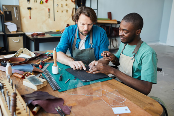 male artist teaching young apprentice using wheelchair in leatherworkers workshop